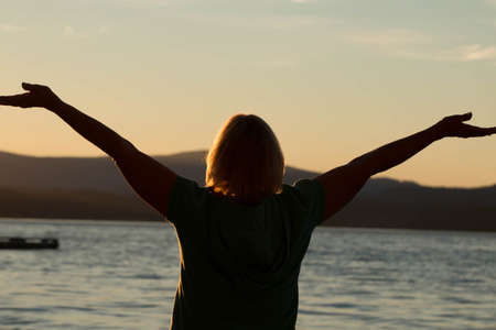 Back of mature woman, standing on the beach facing the ocean and sunset, with arms outstretched.の写真素材