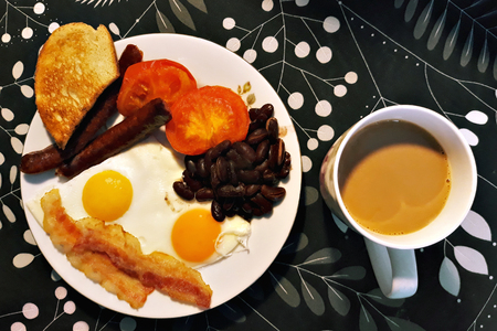 Plate with breakfast consisting of scrambled eggs, ham, sausages and beans and a nearby cup with hot...