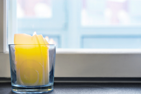 Image of a burning candle standing in a glass on the windowsill against the window.の写真素材