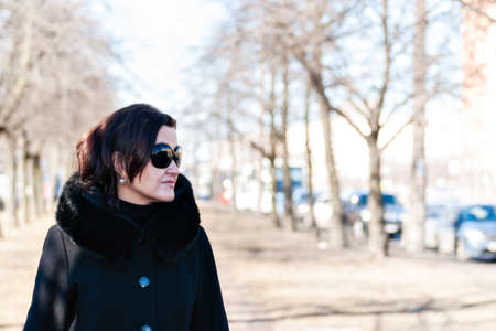 Young woman in a black coat, dark sunglasses and earrings walks along a city street, illuminated by bright spring sunlight.の写真素材