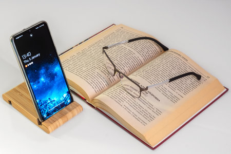 Switched on mobile phone installed on a special stand and an open book with glasses on a white background.の写真素材