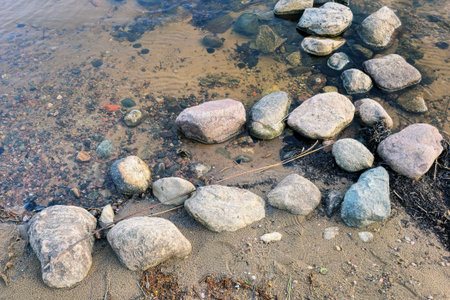 Large and small stones lie on the sand and in clear water on the shore of a lake on a summer day.の写真素材