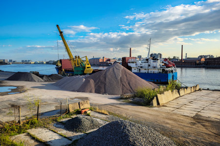 Construction site on the river bank with large piles of rubble, a yellow crane and a barge at the pier on a bright sunny day.の写真素材