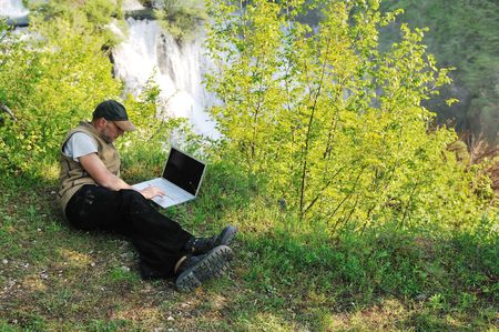 young businessman with hat work on thin laptop outdoor in nature with beautiful waterfalls in background representing freedom and wireless technology concept の写真素材