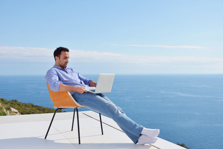 handsome young man relaxing and working on laptop computer at home balcony while looking sunsetの写真素材