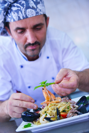 Handsome chef dressed in white uniform decorating pasta salad and seafood fish in modern kitchenの写真素材