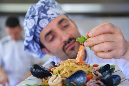 Handsome chef dressed in white uniform decorating pasta salad and seafood fish in modern kitchenの写真素材