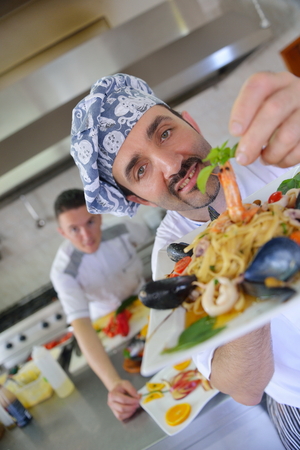 Handsome chef dressed in white uniform decorating pasta salad and seafood fish in modern kitchenの写真素材
