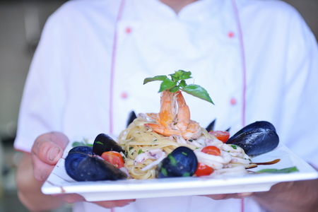 Handsome chef dressed in white uniform decorating pasta salad and seafood fish in modern kitchenの写真素材