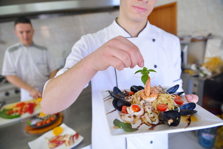 Handsome chef dressed in white uniform decorating pasta salad and seafood fish in modern kitchenの写真素材