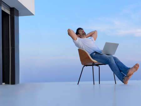 handsome young man relaxing and working on laptop computer at home balcony while looking sunsetの写真素材