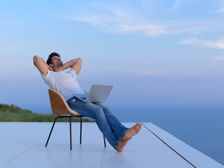 handsome young man relaxing and working on laptop computer at home balcony while looking sunsetの写真素材