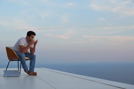 handsome young man relaxing and working on laptop computer at home balcony while looking sunset