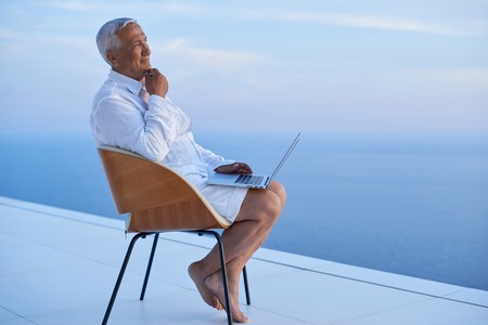 senior man working on laptop computer and looking ocean view sunset on balcony of modern homeの写真素材