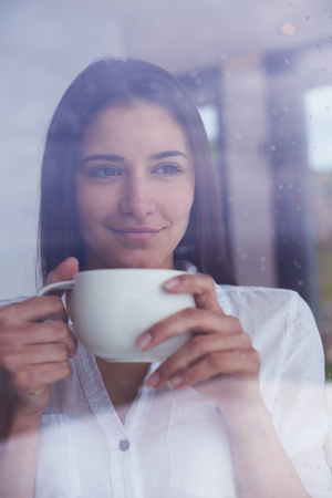 beautiful young woman drink first morning coffee at modern home interior with rain drops on big window door glassの写真素材