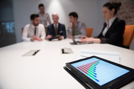 Close up of tablet touchpad computer with focus on business analytics and progress chart document. Business people group on meeting in backgroundの写真素材