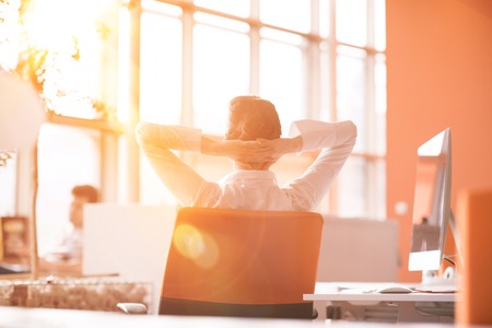 Happy young business woman relaxing and geting insiration while working on desktop computer at modern bright  starup office interior. Morning sunrise or sunset with sun flare in background.の写真素材