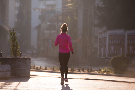 sporty woman running on sidewalk at early morning jogging with city  sunrise scene in backgroundの写真素材