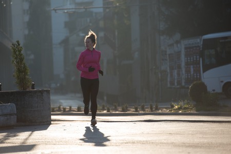 sporty woman running on sidewalk at early morning jogging with city  sunrise scene in backgroundの写真素材