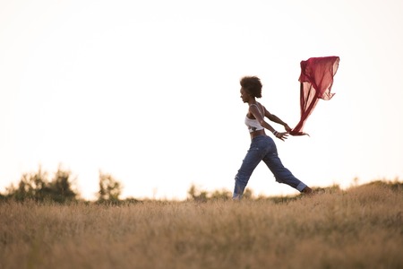 Young beautiful black girl laughs and dances outdoors with a scarf in her hands in a meadow during sunsetの写真素材
