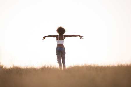 Young beautiful black girl laughs and dances outdoors in a meadow during sunset