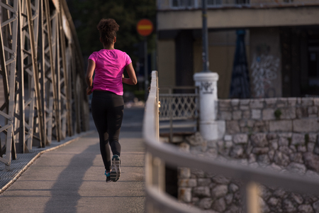 Young sporty african american woman running on sidewalk across the bridge at early morning jogging with city sunrise scene in backgroundの写真素材