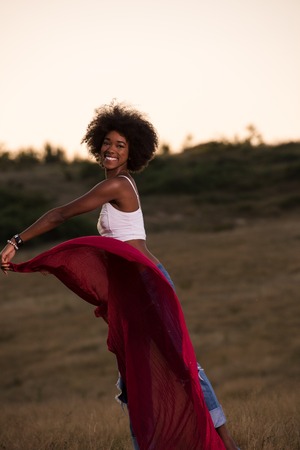 Young beautiful black girl laughs and dances outdoors with a scarf in her hands in a meadow during sunsetの写真素材