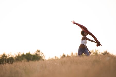 Young beautiful black girl laughs and dances outdoors with a scarf in her hands in a meadow during sunsetの写真素材
