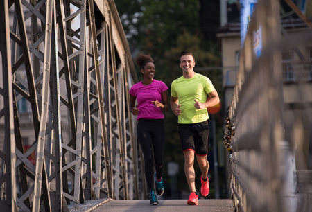 healthy young multiethnic couple jogging across the bridge in the city at early morning with sunrise in backgroundの写真素材