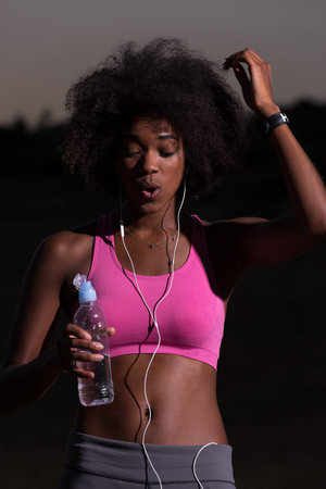 portrait of a young beautiful african american woman with headphones and a bottle of water after jogging in nature beautiful summer nightの写真素材
