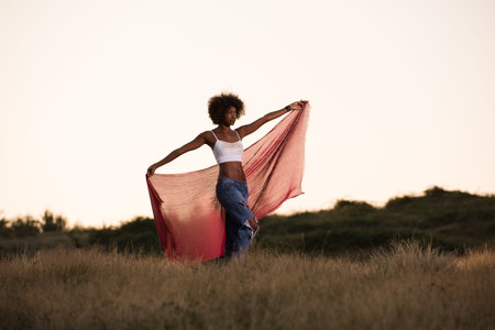 Young beautiful black girl laughs and dances outdoors with a scarf in her hands in a meadow during sunsetの写真素材