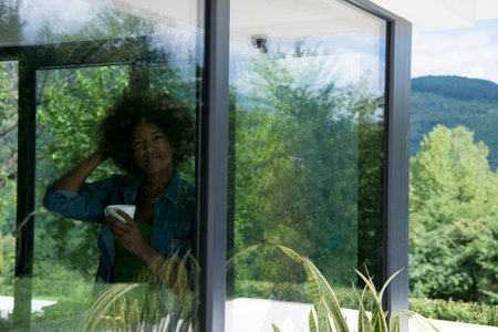 Beautiful young african american woman drinking coffee and looking through a window in her luxury...
