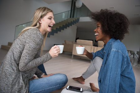Two young laughing multiethnic women sit on the floor and enjoy while drinking coffee using smartphone in luxury homeの写真素材
