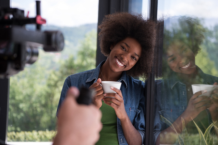 Beautiful young african american woman drinking coffee and looking through a window in her luxury homeの写真素材