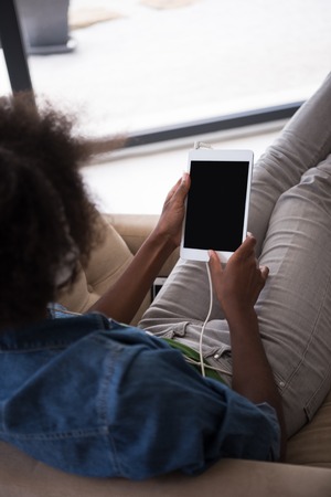 young beautiful African American woman enjoys listening to music with headphones and tablet in your armchair in her luxury homeの写真素材