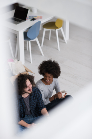Relaxing in new house. Cheerful young multiethnic couple sitting on the floor and drinking coffee while cardboard boxes laying all around themの写真素材