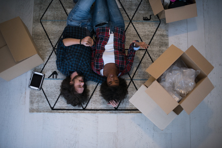 Top view of attractive young multiethnic couple moving, holding hands, looking at camera and smiling while lying among cardboard boxesの写真素材