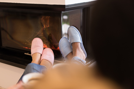 Young romantic multiethnic couple sitting on sofa in front of fireplace at home, looking at each other, talking and drinking coffeeの写真素材