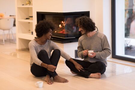 Young romantic multiethnic couple sitting on the floor in front of fireplace at home, looking at each other, talking and drinking coffee autumn dayの写真素材