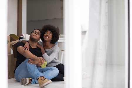 Relaxing in new house. Cheerful young African American couple sitting on the floor and drinking coffee while cardboard boxes laying all around themの写真素材