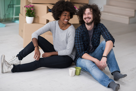 Relaxing in new house. Cheerful young multiethnic couple sitting on the floor and drinking coffee while cardboard boxes laying all around themの写真素材