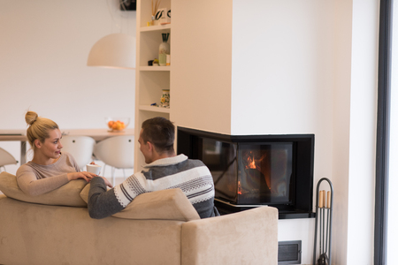 Young romantic couple sitting on sofa in front of fireplace at home, looking at each other, talking and drinking coffee autumn dayの写真素材