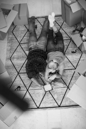 Top view of attractive young couple moving, holding hands, looking at camera and smiling while lying among cardboard boxesの写真素材