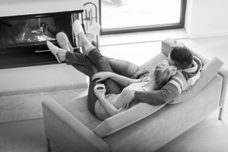 Young romantic couple sitting on sofa in front of fireplace at home, looking at each other, talking and drinking coffee autumn dayの写真素材