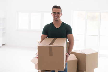 Moving to a new apartment. Cheerful young man holding a cardboard boxes and smiling at camera while other carton boxes laying on backgroundの写真素材