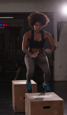 Fit young african american woman box jumping at a crossfit style gym. Female athlete is performing box jumps at gym.の写真素材