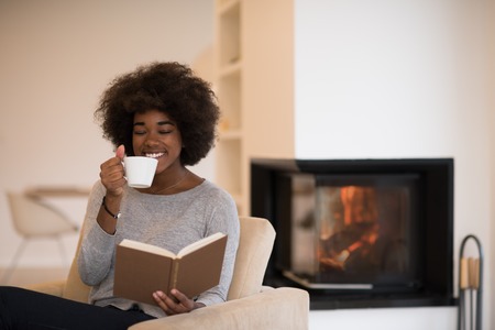 african american woman drinking cup of coffee reading book at fireplace. Young black girl with hot beverage relaxing heating warming up. autumn at home.の写真素材