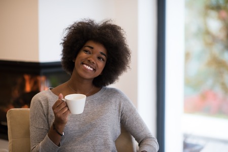 Happy joyful african american woman drinking cup of coffee relaxing at fireplace. Young black girl with hot beverage heating warming up. autumn at home.の写真素材