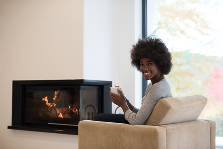 Happy joyful african american woman drinking cup of coffee relaxing at fireplace. Young black girl with hot beverage heating warming up. autumn at home.の写真素材
