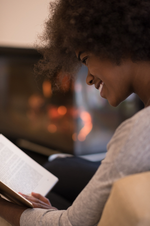 Young beautiful african american woman sitting in front of fireplace at home on a cold autumn day and reading bookの写真素材